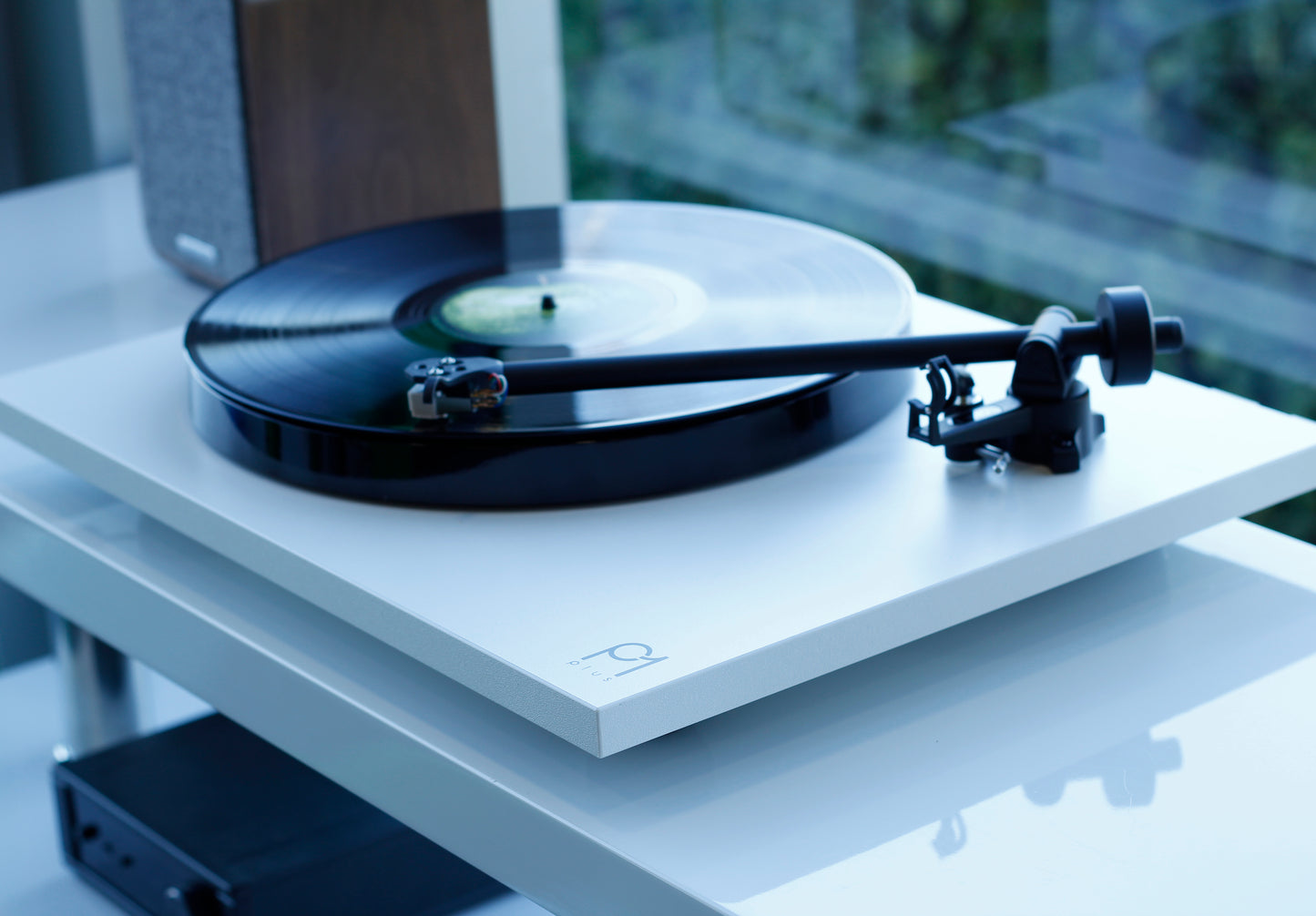 Product image for a Rega Planar 1 Plus turntable in Matt White finish next to a window. The shelf below shows a Rega io Amplifier